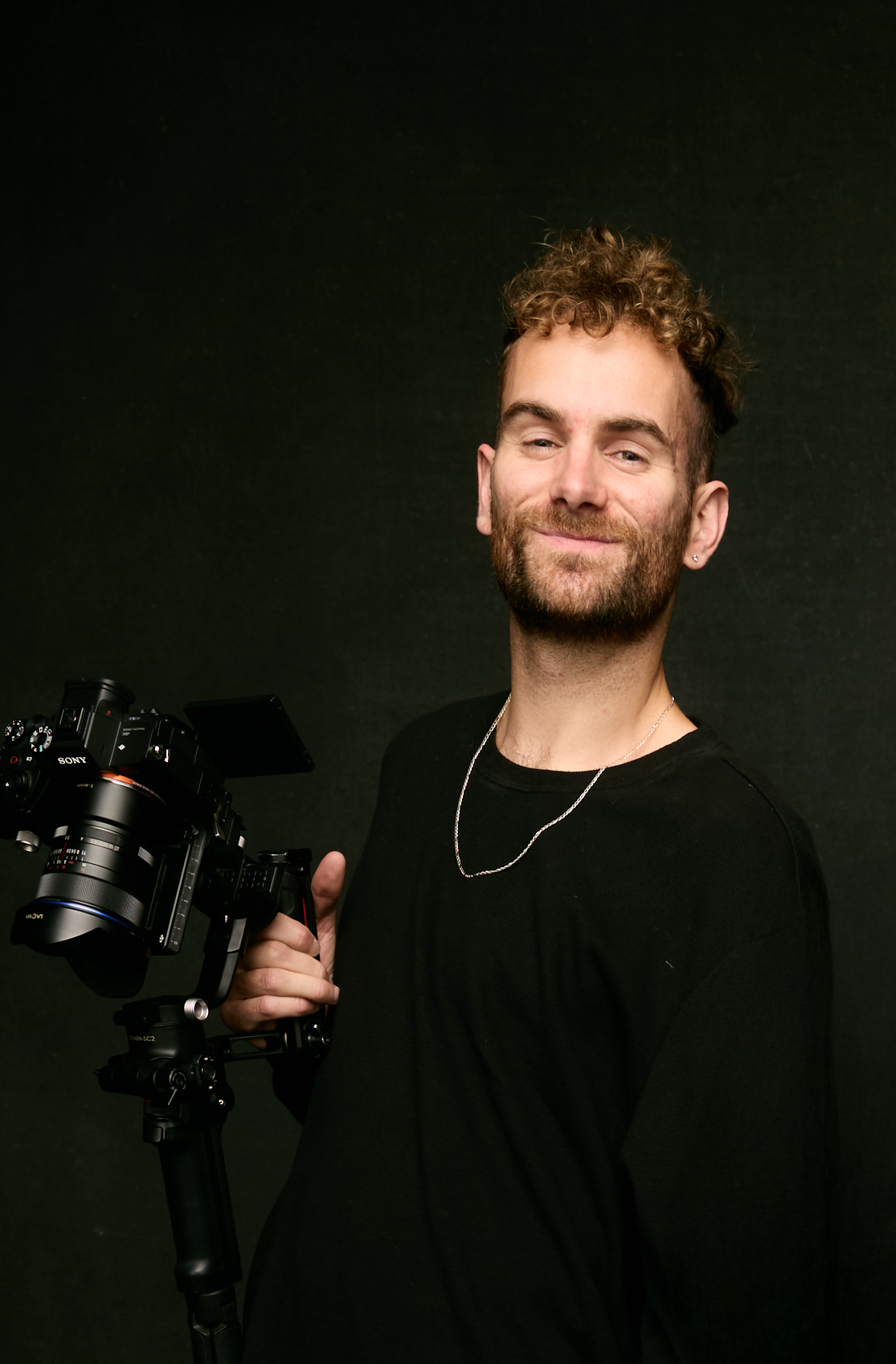 A young man with curly hair and a beard smiling while holding a professional camera on a gimbal.
