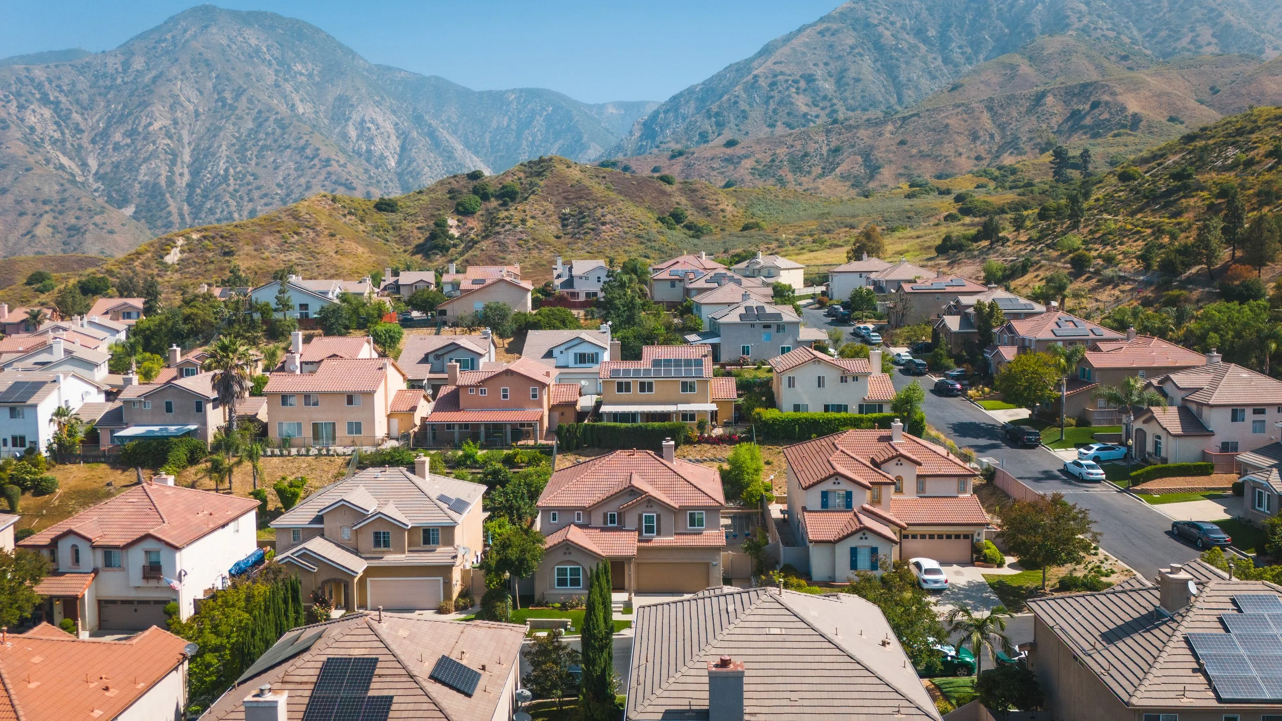 Aerial view of a suburban neighborhood with houses, trees, and streets, set against a mountainous backdrop.