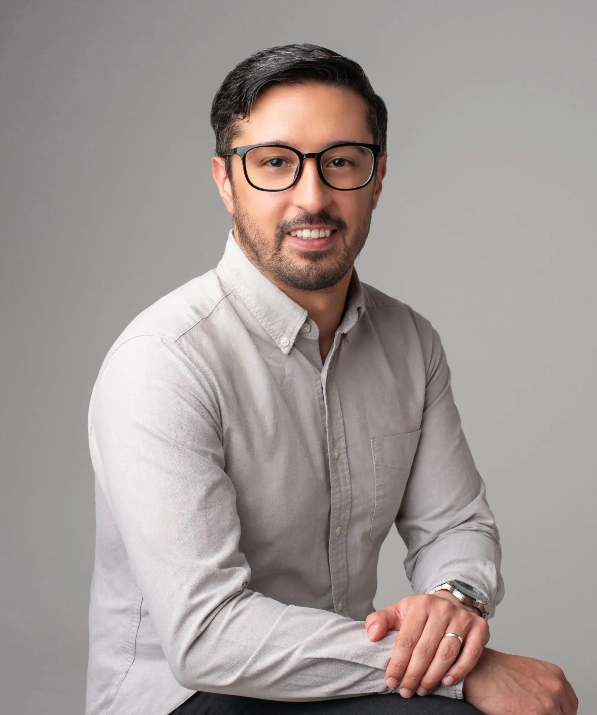 A man with dark hair, beard, and glasses wearing a light grey button-up shirt, smiling, against a plain grey background.