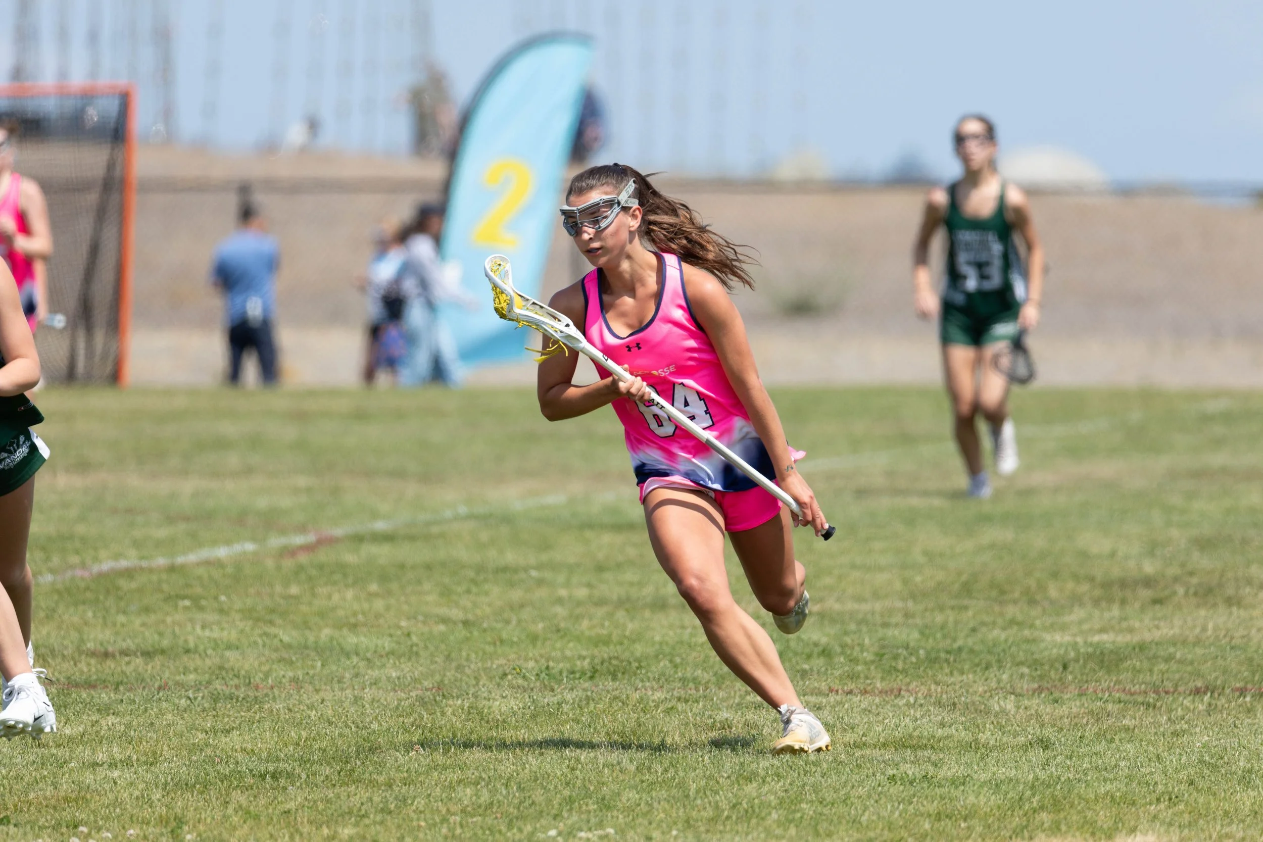 A woman running on a grassy field while holding a lacrosse stick during a sports game. She is wearing a pink sports uniform, goggles, and cleats. Other players and spectators are visible in the background.