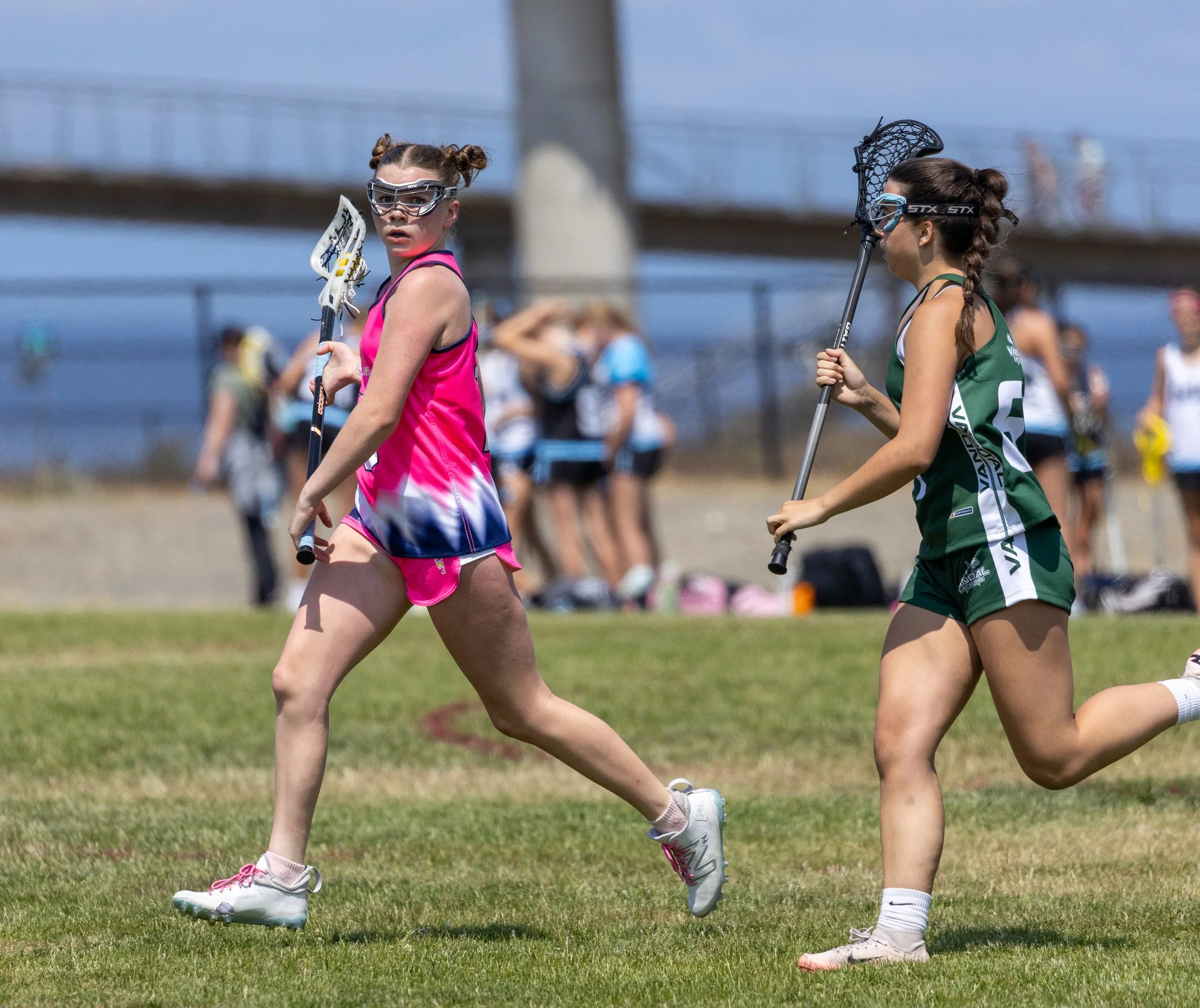 Two young women playing lacrosse outdoors on a grassy field, with one in a pink and blue uniform and the other in a green and white uniform, both holding lacrosse sticks and wearing goggles.