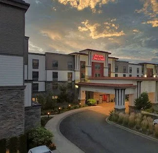 A modern hotel building with a curved driveway leading to the entrance, set against a sunset sky with scattered clouds.