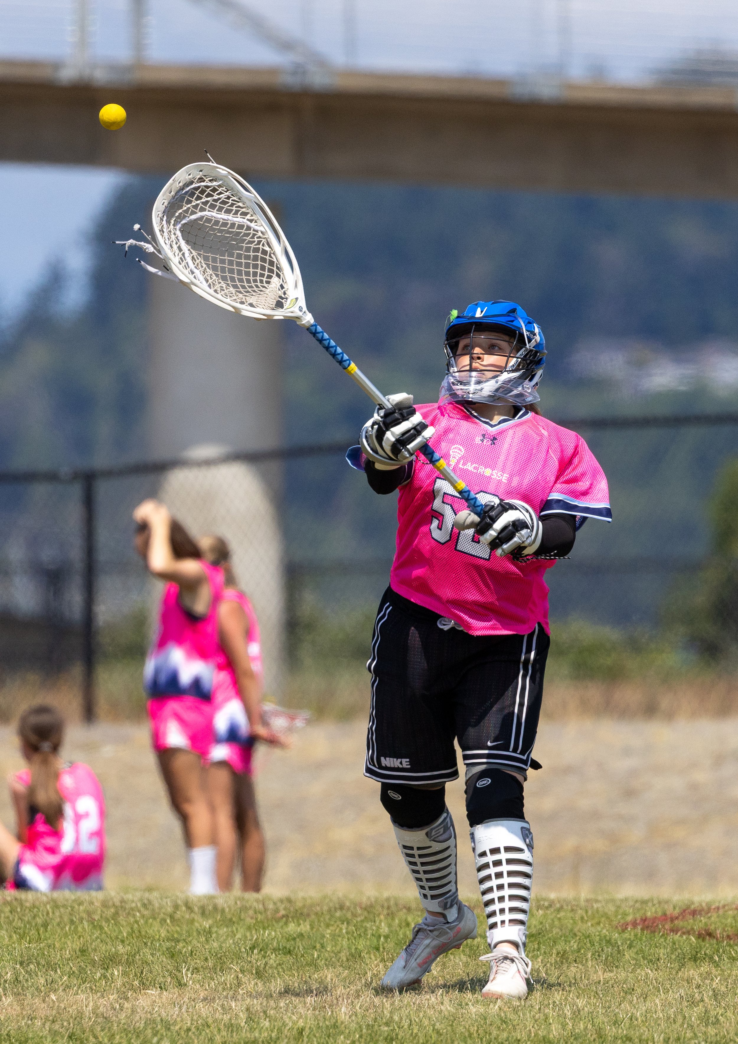 A girl in a pink sports jersey with the number 52, wearing protective gear and a helmet, plays lacrosse outdoors on a grassy field, reaching out with her stick to catch a yellow lacrosse ball.