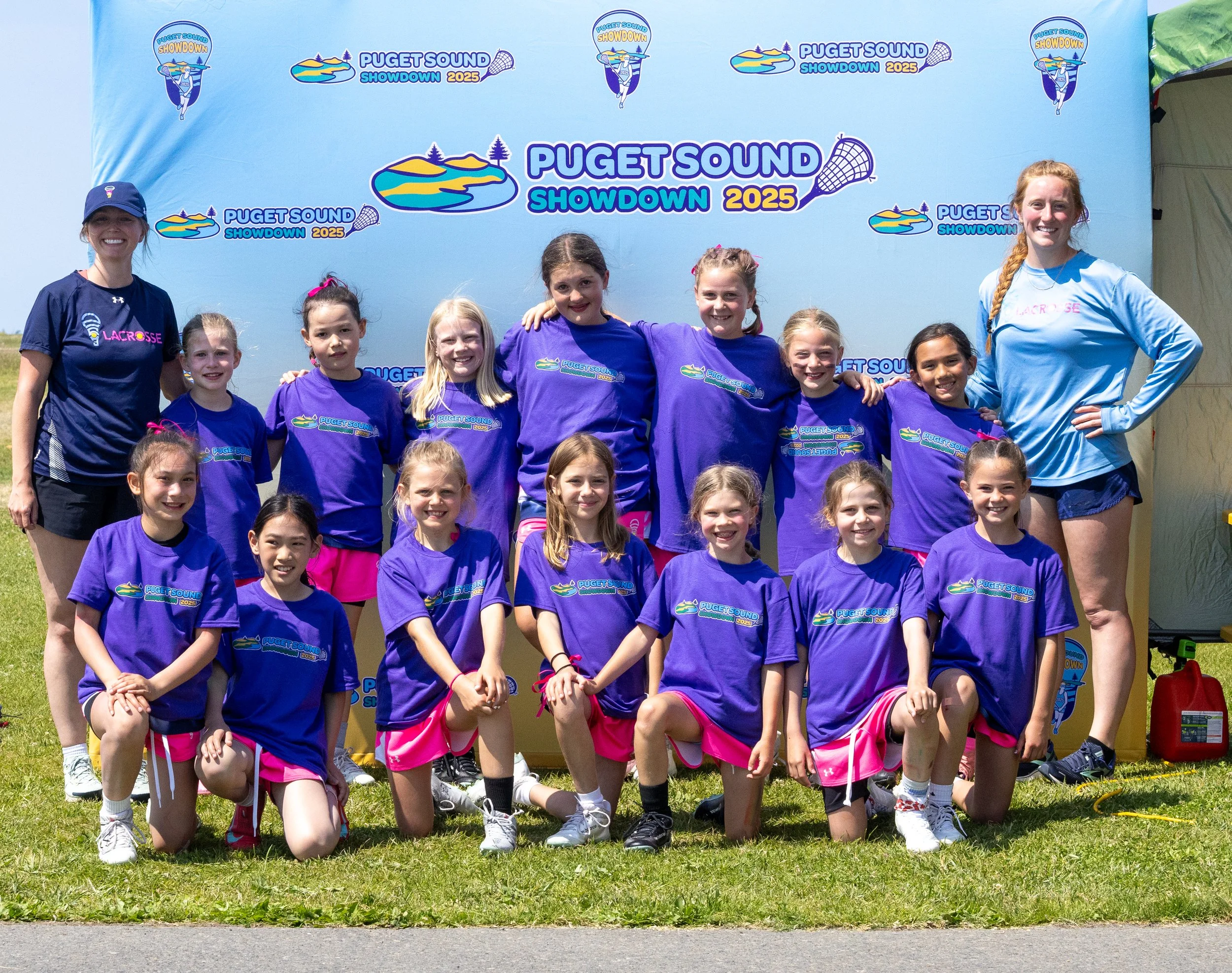 A group of young girls and two coaches posing for a photo at a sports event called 'Puget Sound Showdown 2025.' The girls are wearing purple shirts with the event logo, some with pink shorts, and the coaches are dressed in sportswear. They are standing outdoors in front of a blue backdrop with the event name and logo.