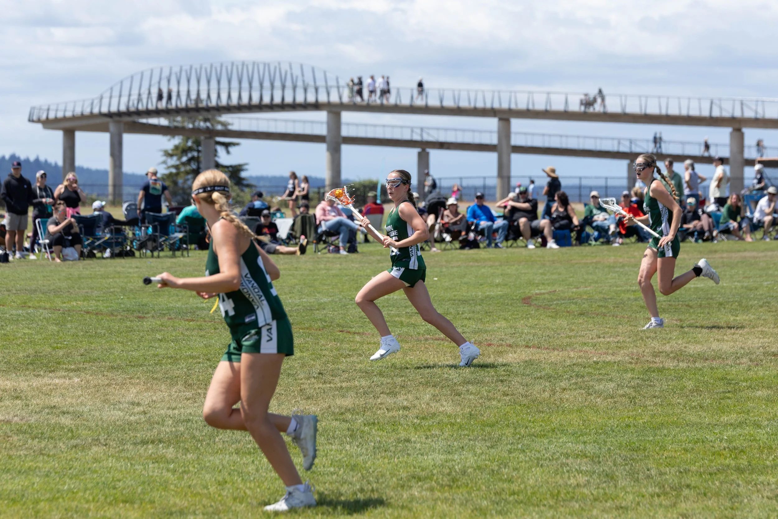 Girls playing lacrosse on a grassy field with spectators sitting and standing in the background, and a modern lookout structure in the distance.