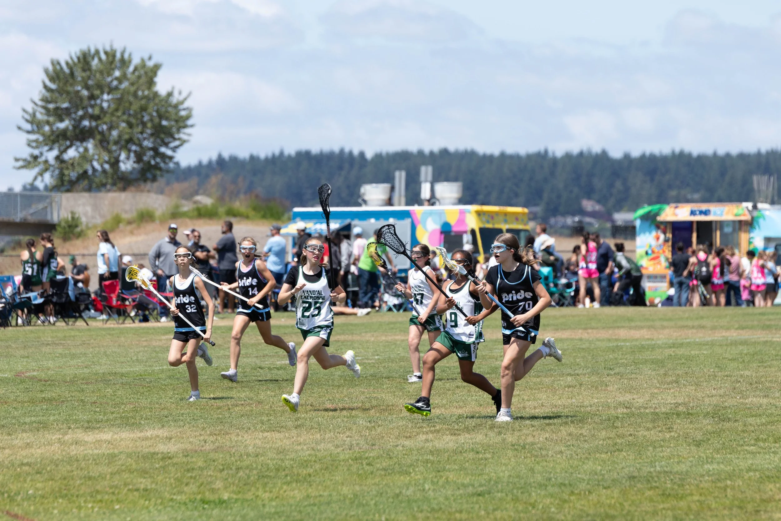 Young girls playing lacrosse on a field during a sports event with spectators and food trucks in the background.