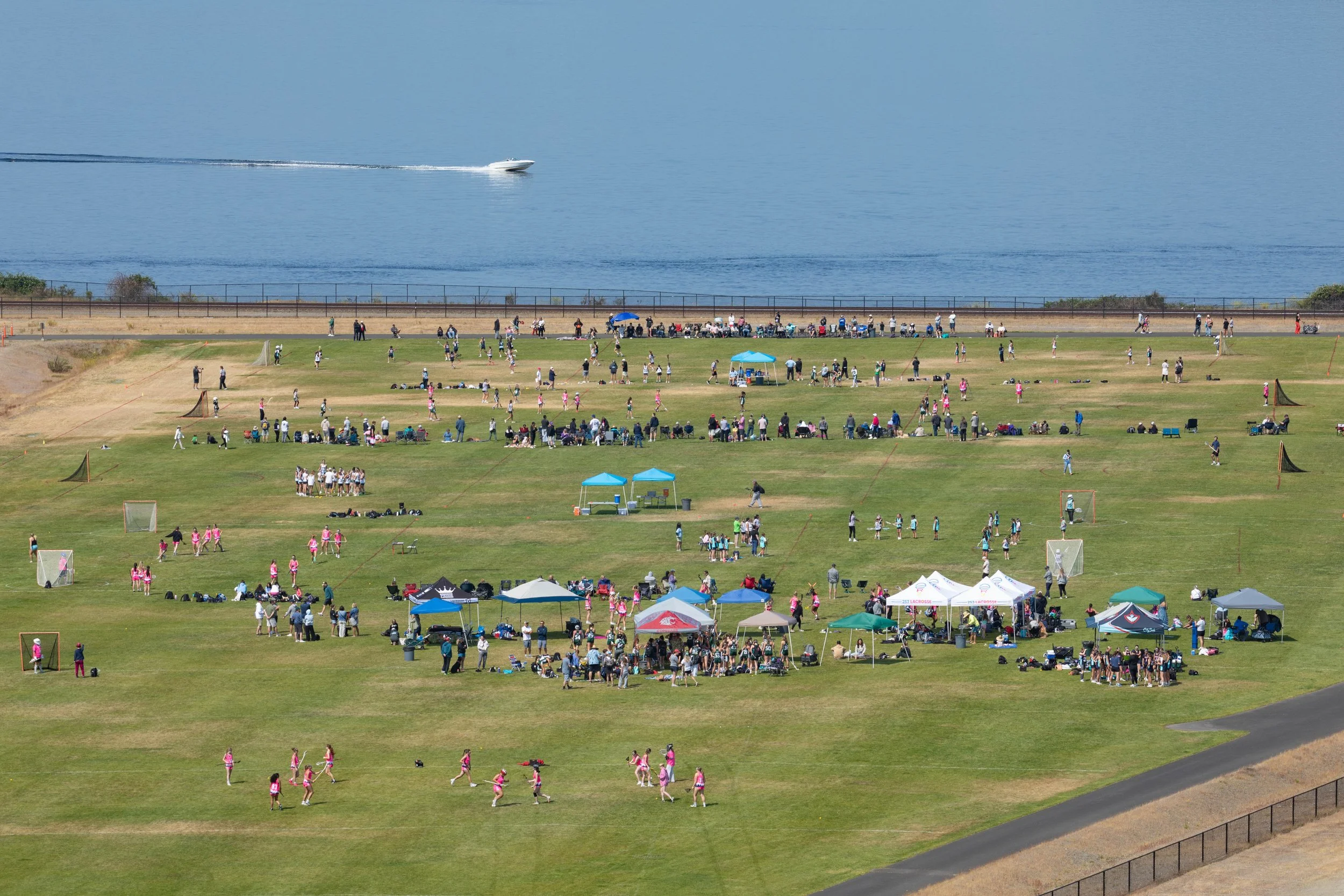 A large outdoor field near water with people participating in sports and viewing activities. Tents and groups of spectators are set up on the grass, and the scene includes a body of water with a speedboat in motion in the background.
