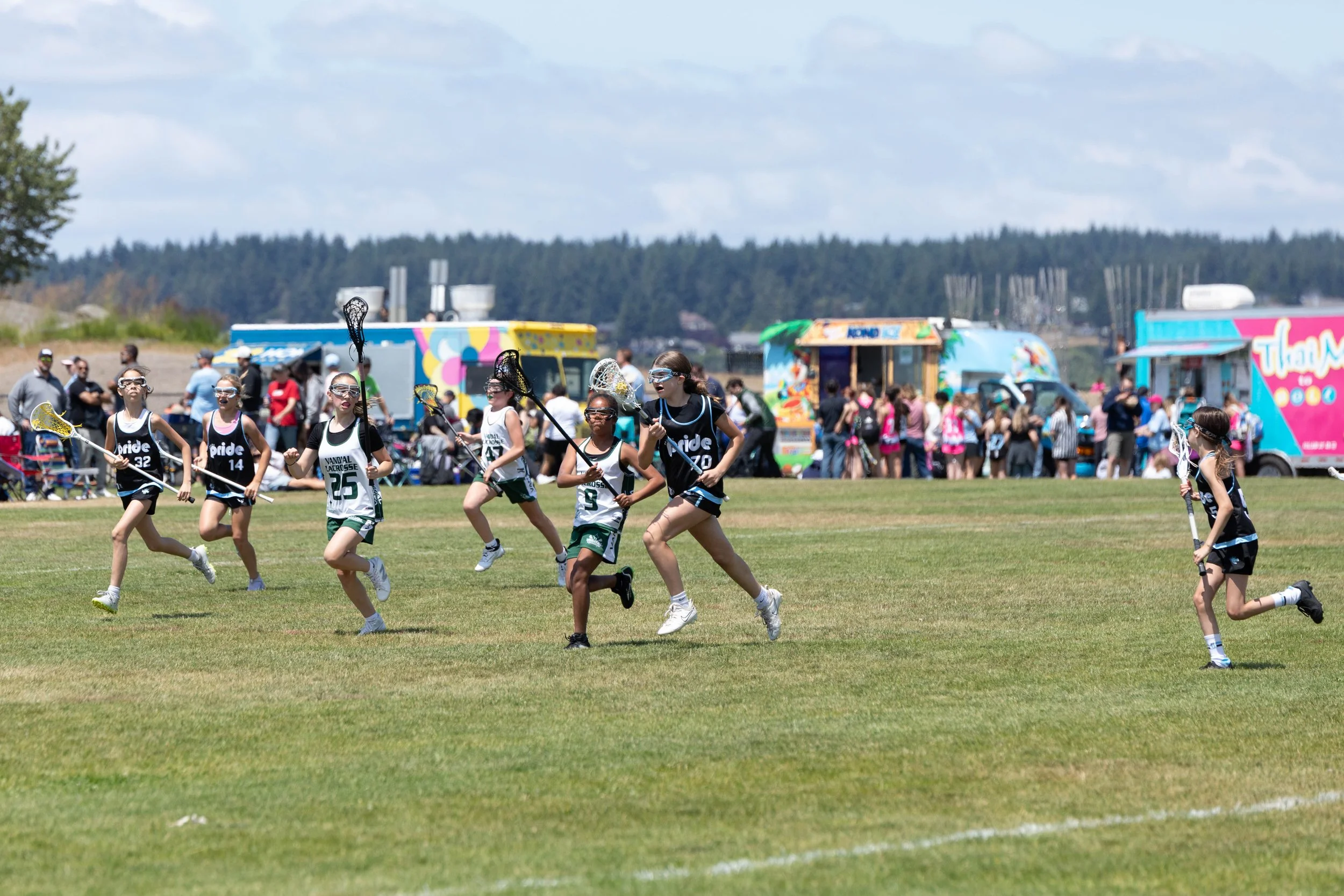 Group of young girls playing lacrosse on a grassy field during a sunny day with food trucks and spectators in the background.