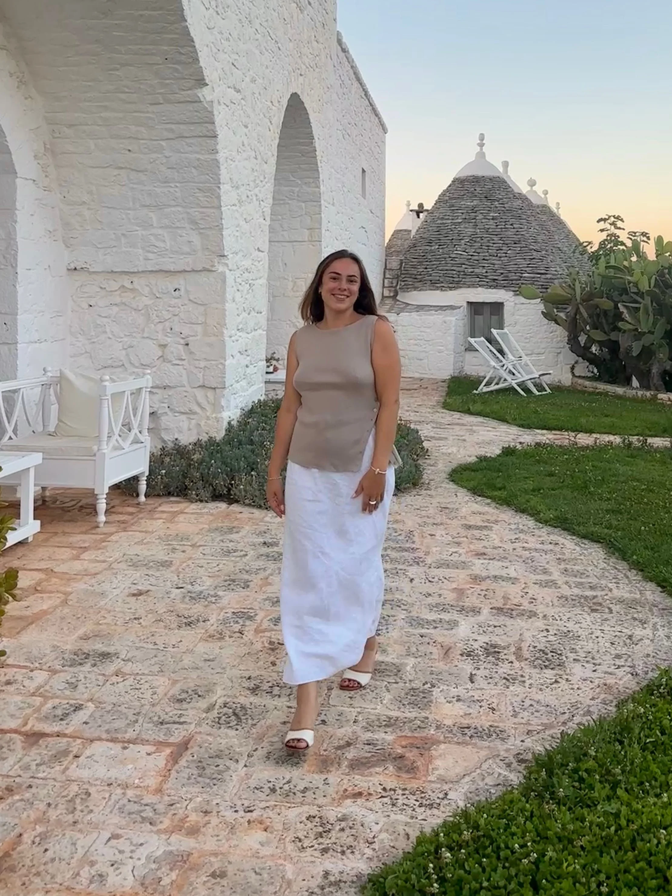 A woman in a beige sleeveless top, white skirt, and high heels standing on a stone-paved path outdoors.
