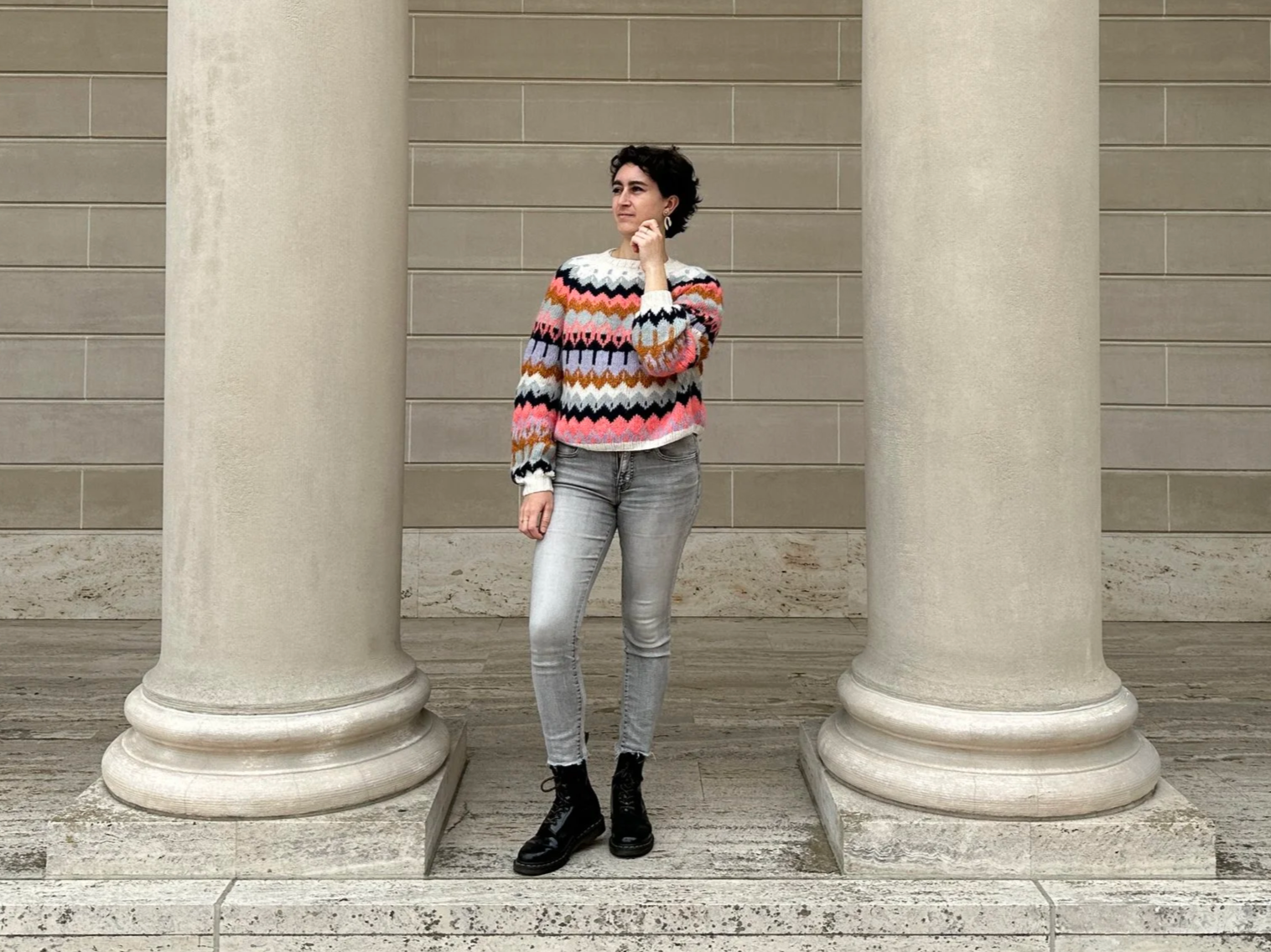 Woman standing between two large columns in front of a beige stone wall, wearing a colorful zigzag patterned sweater, grey jeans, and black boots.