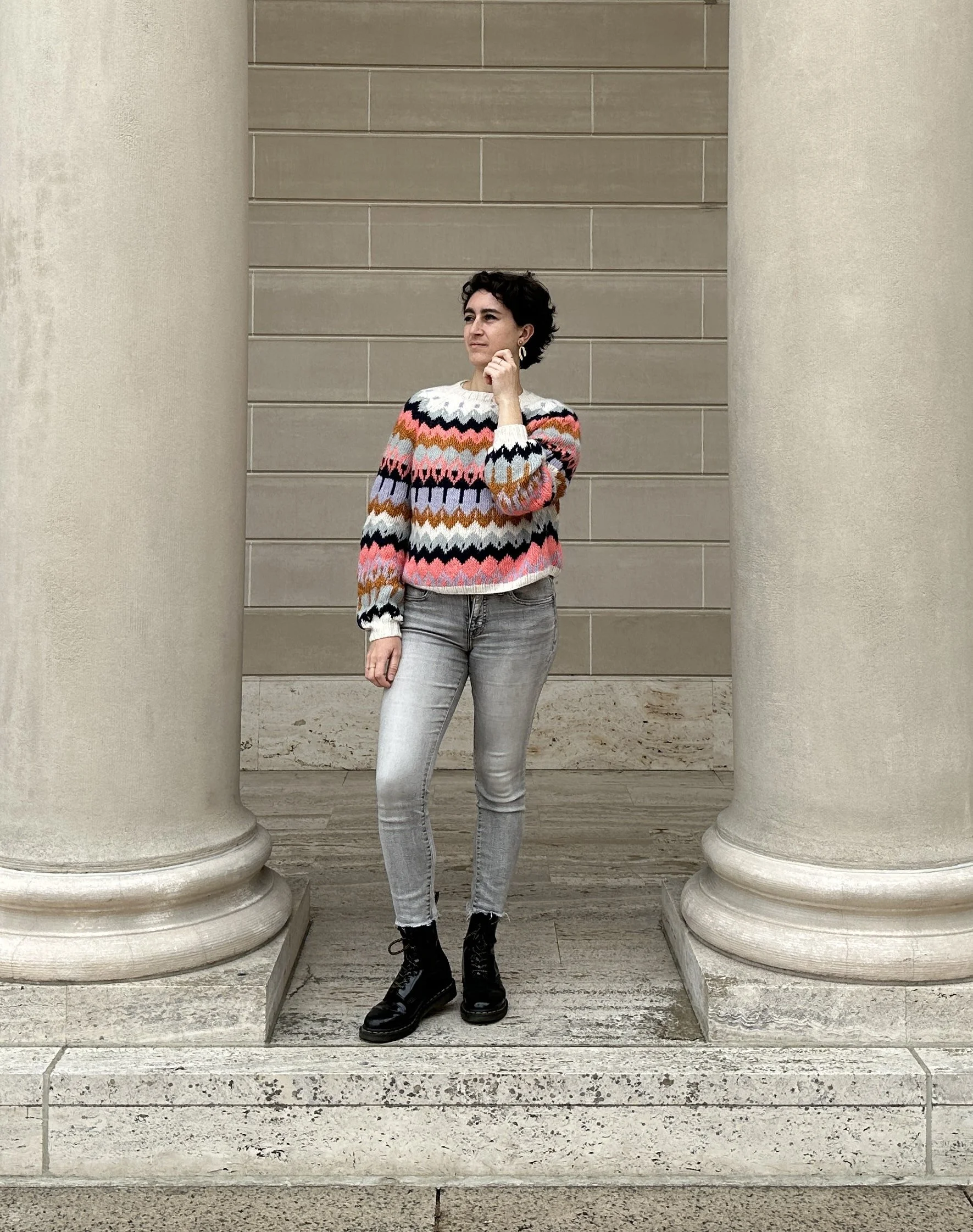 Woman standing between two large columns in front of a beige stone wall, wearing a colorful zigzag patterned sweater, grey jeans, and black boots.