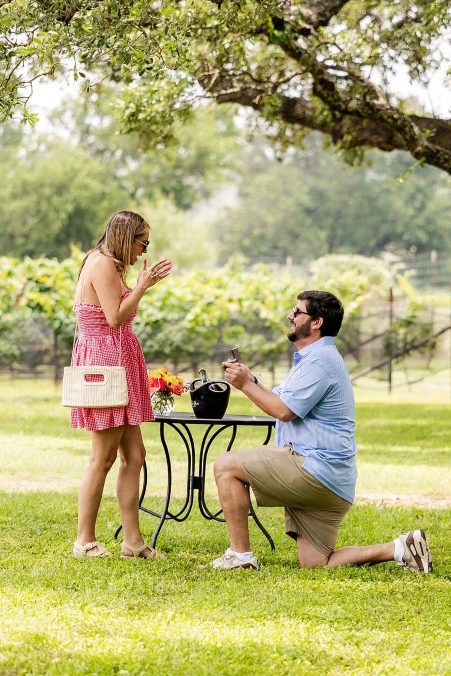 A man is proposing marriage to a woman in a park, with the woman appearing surprised and happy. The man is kneeling on one knee and holding a small box, possibly with an engagement ring, while the woman is holding her hands to her face. They are outdoors on a grassy area with a tree overhead and a small table with flowers and a gift bag between them.
