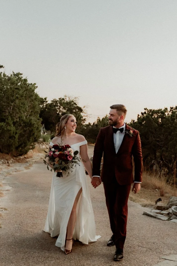 A newlywed couple walking hand in hand on a pathway outdoors at sunset, with the bride holding a bouquet of flowers and wearing a white wedding gown, and the groom in a dark red suit with a black bow tie.