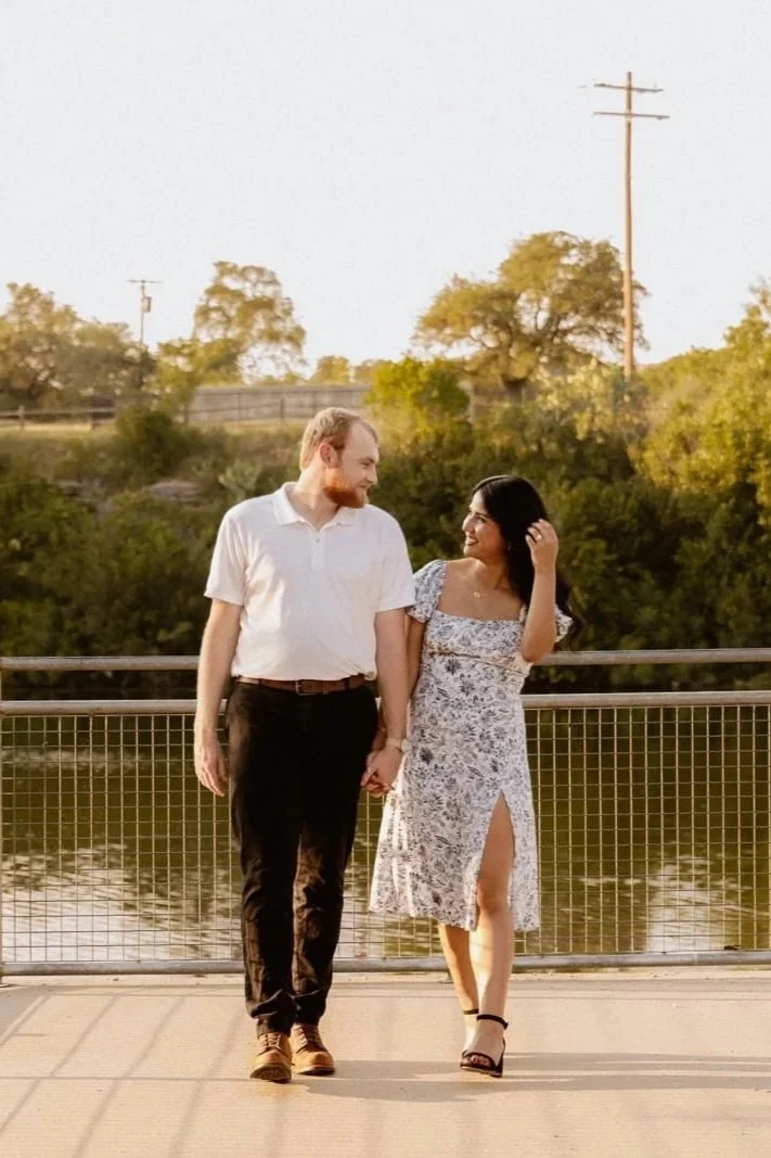 A couple holding hands and walking on a bridge near a body of water, with trees and power lines in the background, during sunset.