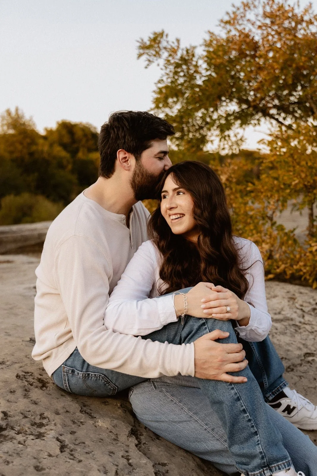 A happy couple sitting on a rock outdoors during fall, with trees with orange leaves in the background. The man is kissing the woman's forehead, and they are smiling, holding hands.