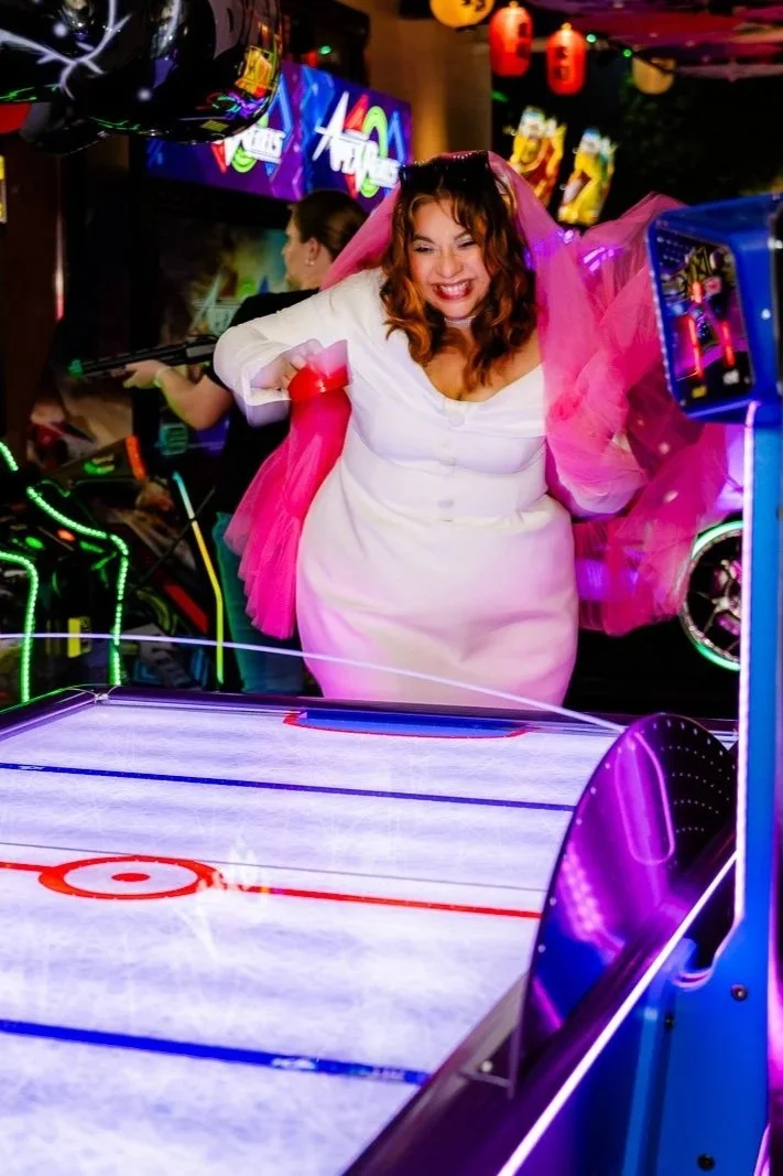 A woman in a white dress with pink tulle celebrating at an arcade, playing air hockey. She is smiling and appears excited, with neon lights and other arcade games visible in the background.