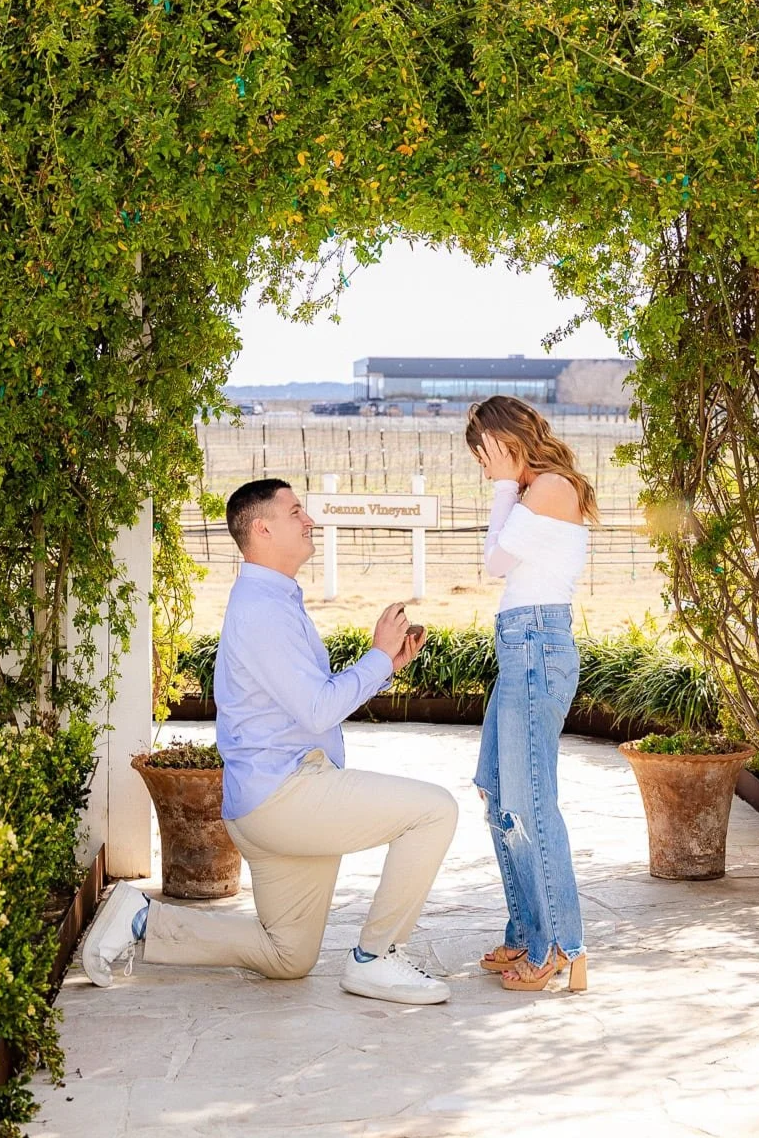 A man proposes marriage to a woman outdoors under a green leafy archway, with a vineyard and a sign reading 'Jocena Vineyard' in the background. The man is kneeling on one knee, holding a ring box, while the woman covers her face with her hands in surprise.
