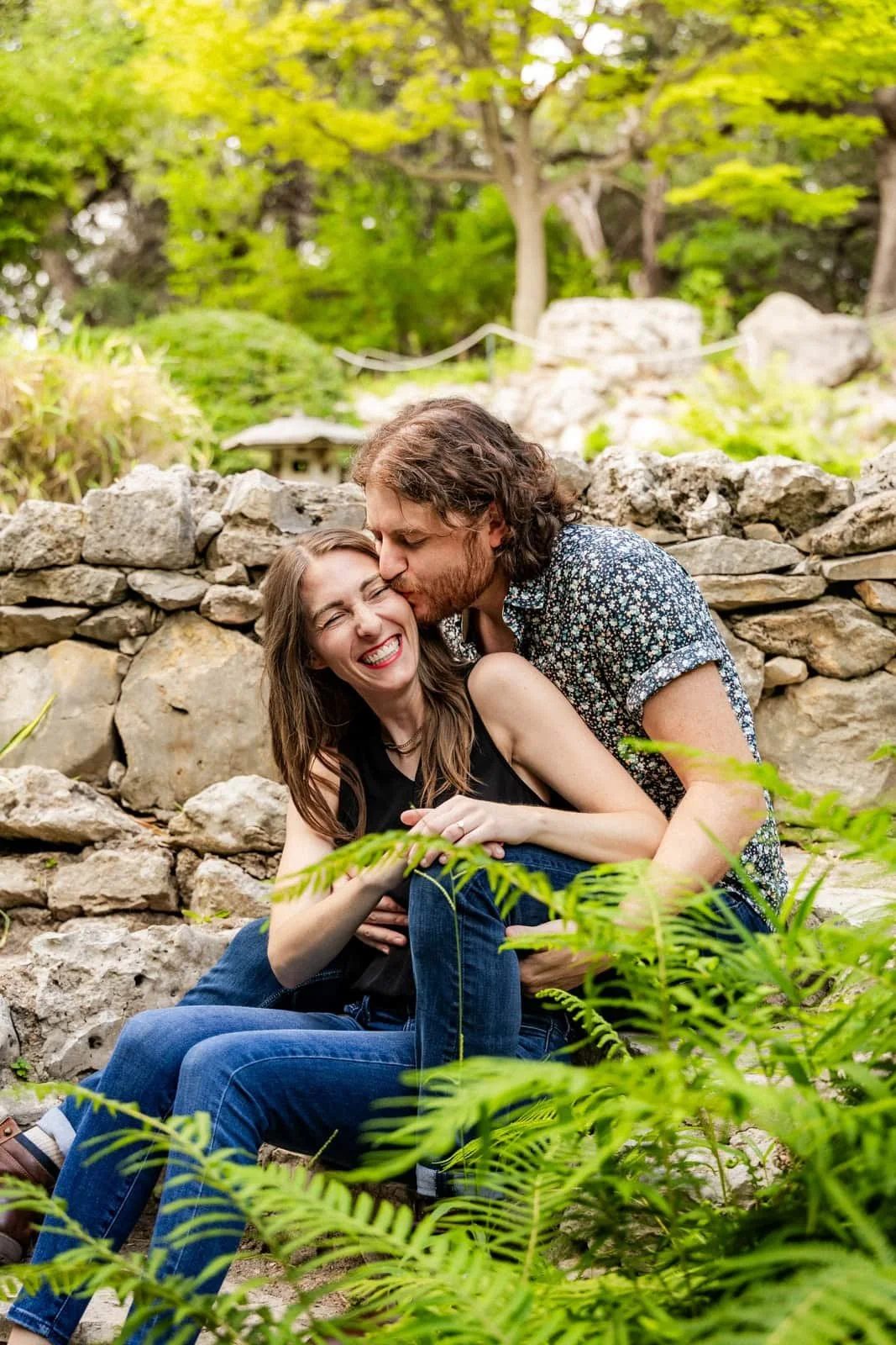 A man kisses a woman on the cheek while sitting on rocks in a lush garden with green foliage.