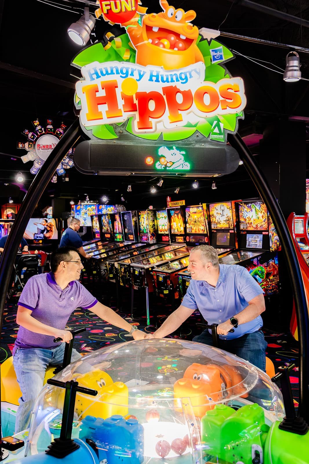 Two men playing an arcade game called 'Hungry Hungry Hippos' at an arcade. The game features colorful hippo figures, with the one on the left being yellow and the one on the right orange. They are holding the game handles and appear to be in motion, smiling and smiling at each other.
