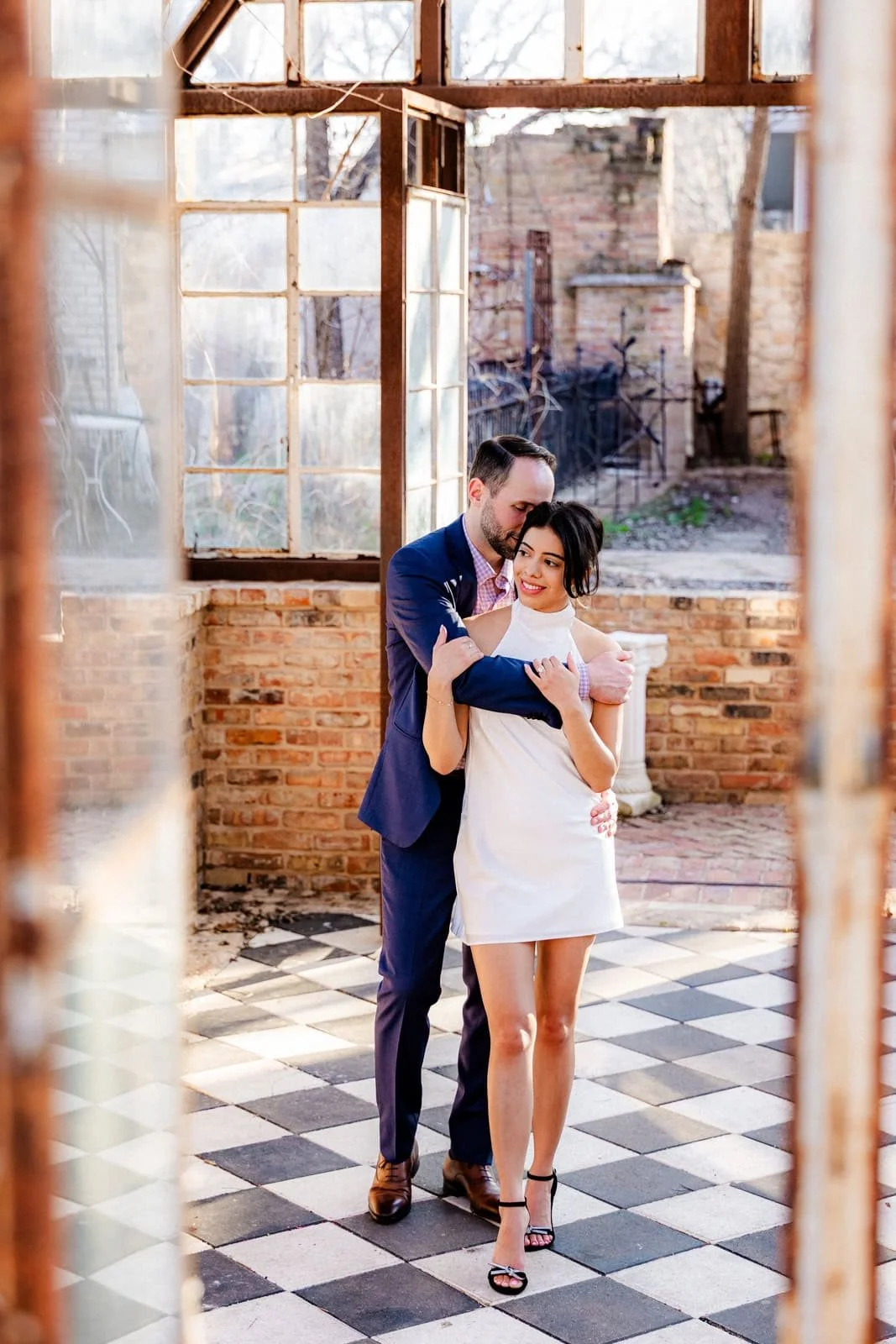 A man and woman in formal attire embracing each other inside a room with large windows and checkered tile floor, brick walls, and outdoor scenery visible through the windows.