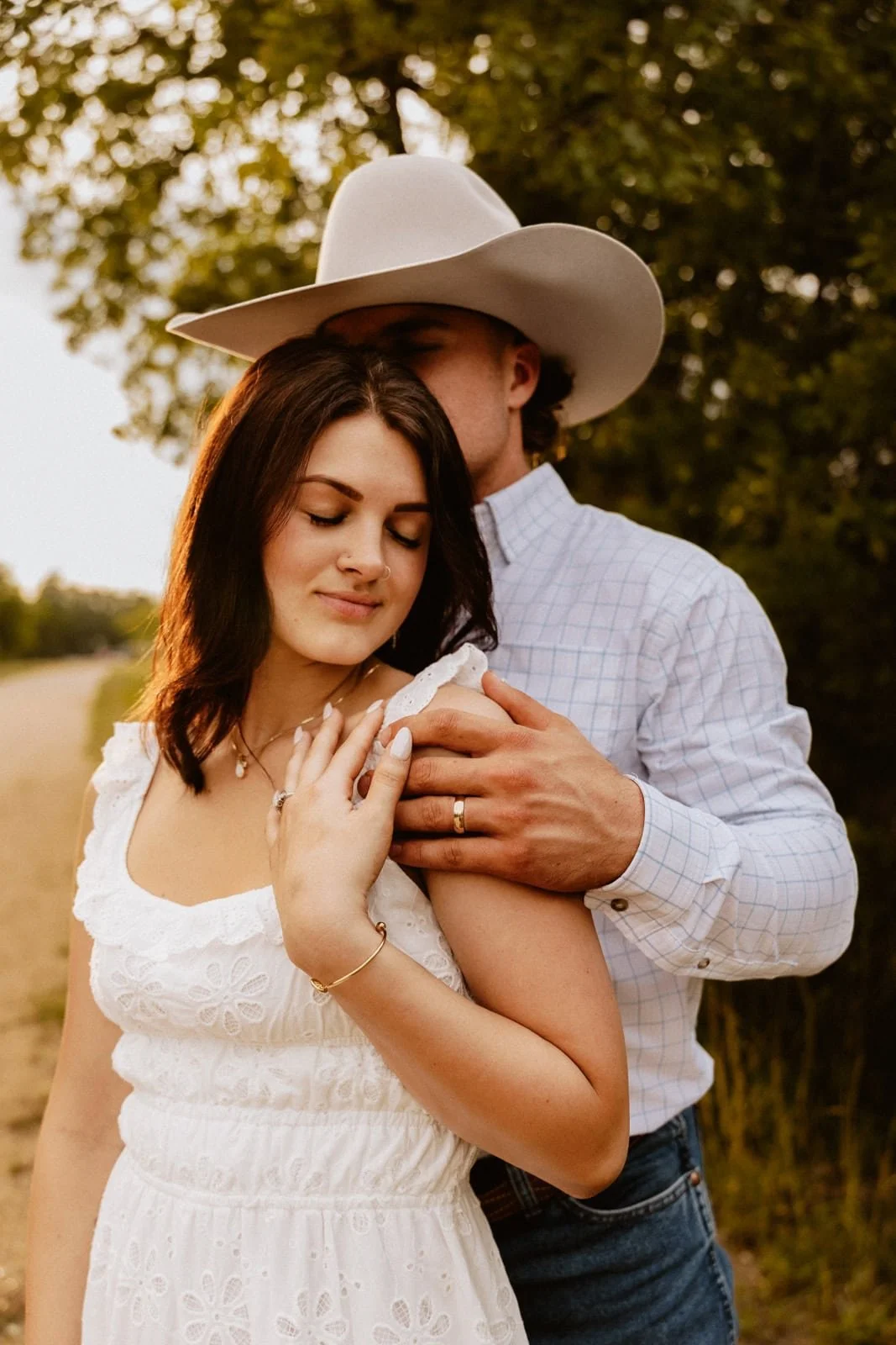 A couple embracing outdoors; the woman with dark hair in a white dress with eyelet pattern and the man wearing a cowboy hat and plaid shirt, standing close with the man's hand on the woman's shoulder, both with serene facial expressions.