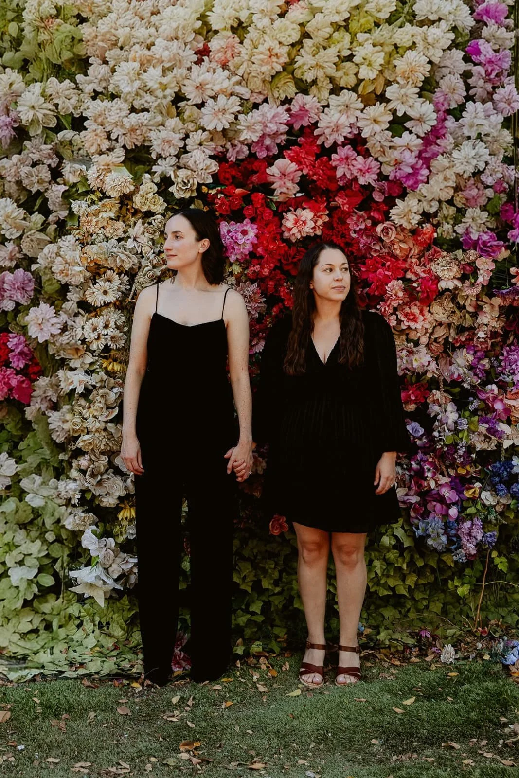 Two women in black dresses holding hands, standing in front of a colorful floral wall.