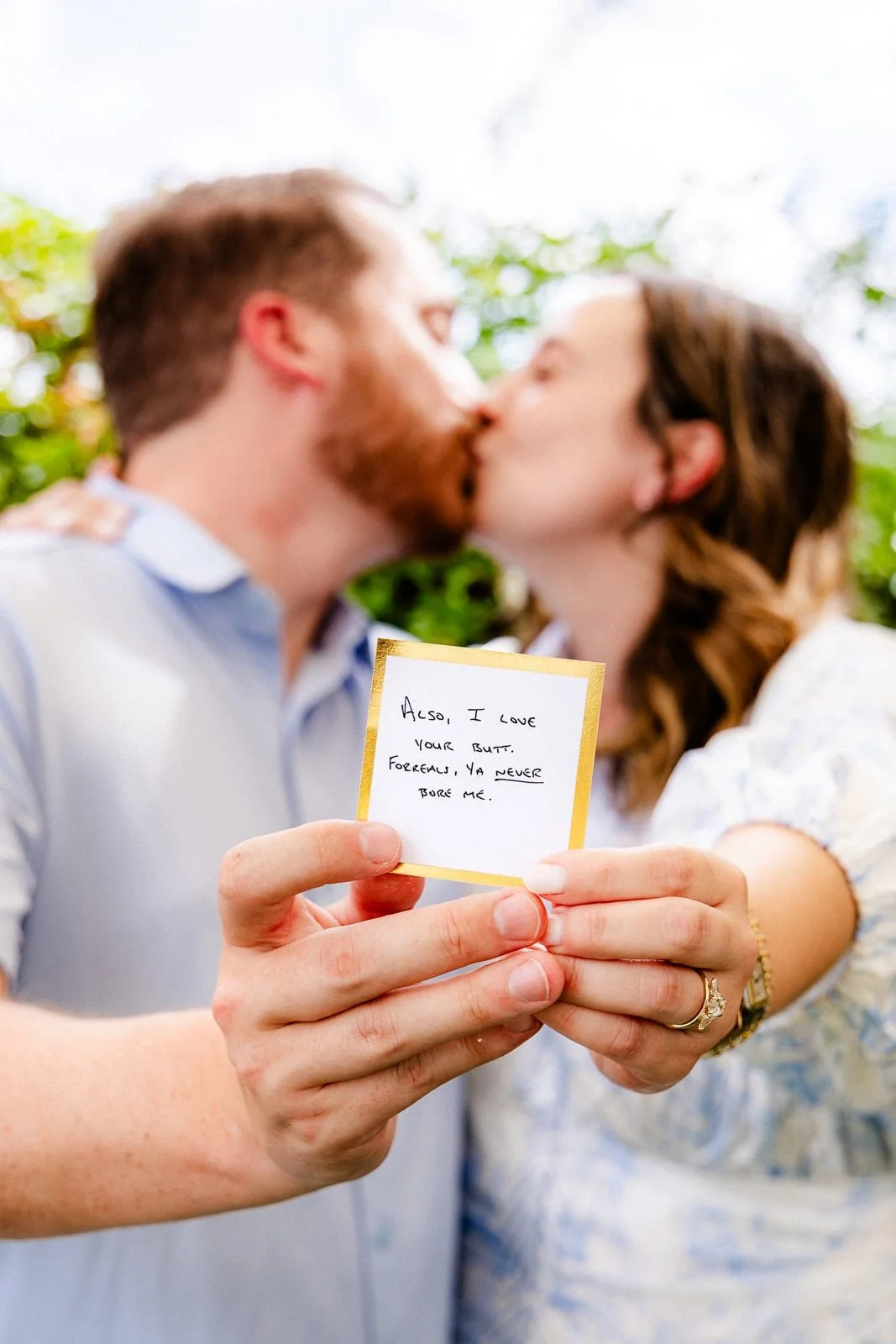 A couple is kissing outdoors, holding a small card with a handwritten message. The man has a beard and is wearing a light blue shirt. The woman has brown hair and is wearing a white patterned top. The card reads: 'Also, I love your butt. Forever, ya never bore me.'