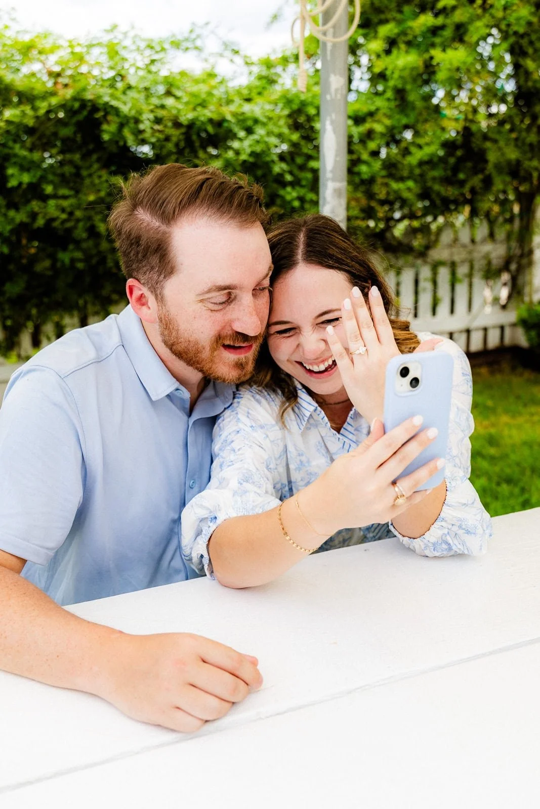 A couple takes a selfie outdoors while sitting at a white table. They are smiling and showing their rings, with a green hedge and a fence in the background.