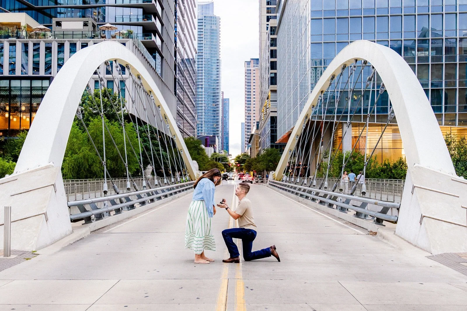 A man proposing marriage to a woman on a city street with modern tall buildings and a white bridge in the background.