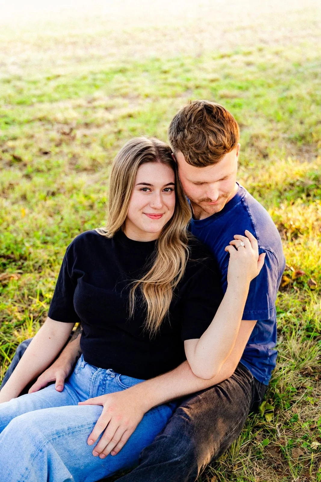 A young couple sitting on the grass in a park, with the woman smiling and leaning on the man's shoulder during sunset.