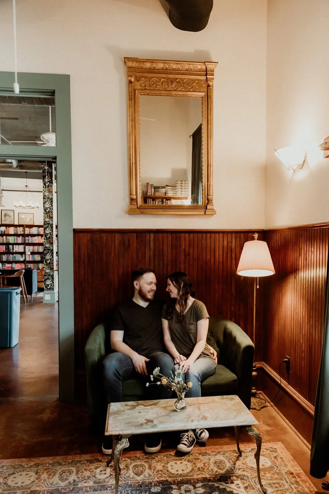 A couple sitting on a green vintage sofa, holding hands, in a cozy room with wooden paneled walls and a lamp beside them. A marble coffee table with a small flower arrangement is in front of them.