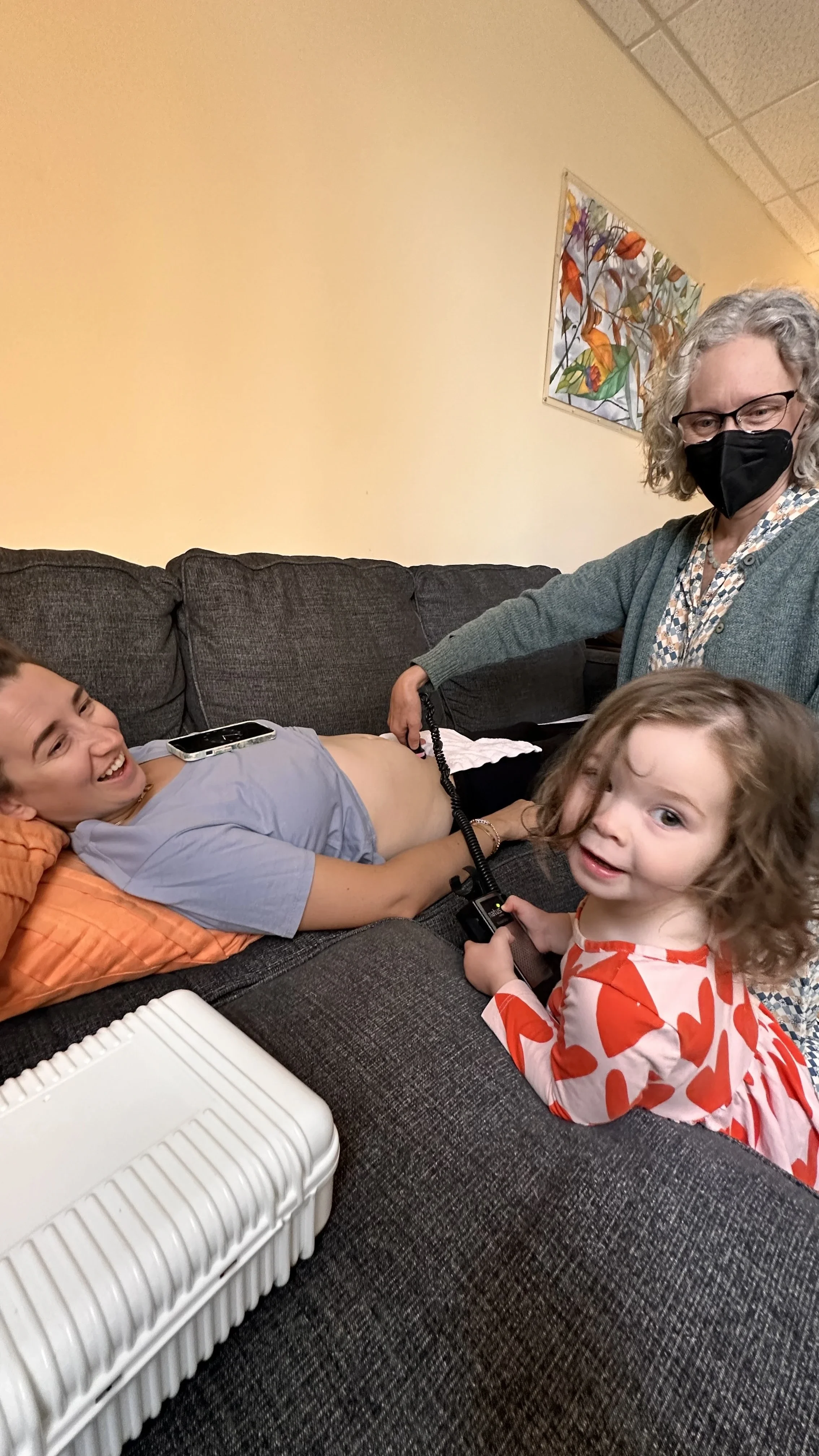 A person lying on a couch having their abdomen checked with a device by a woman, with a young girl sitting on the couch holding the device.
