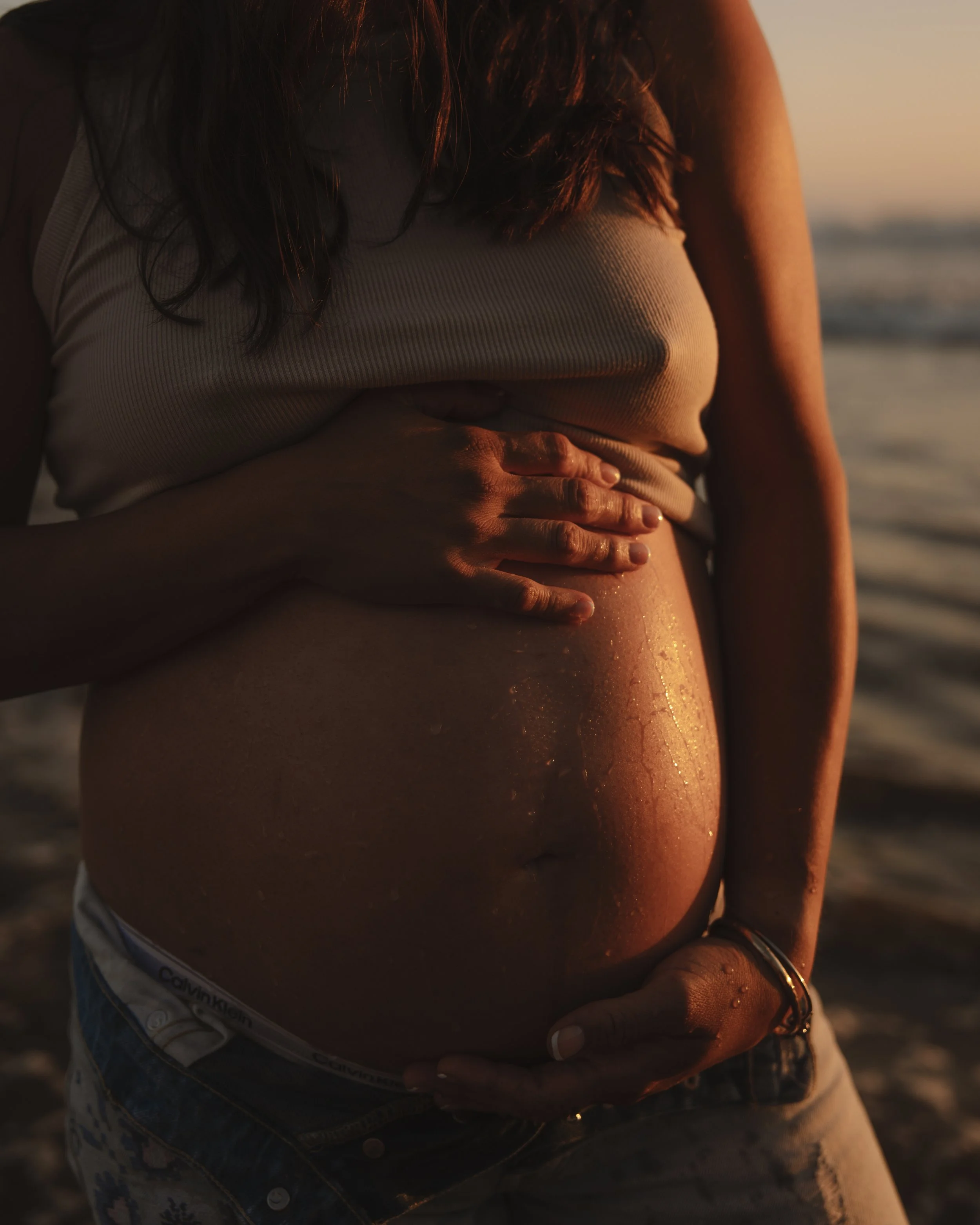 A pregnant woman standing on the beach at sunset, holding her belly with both hands.