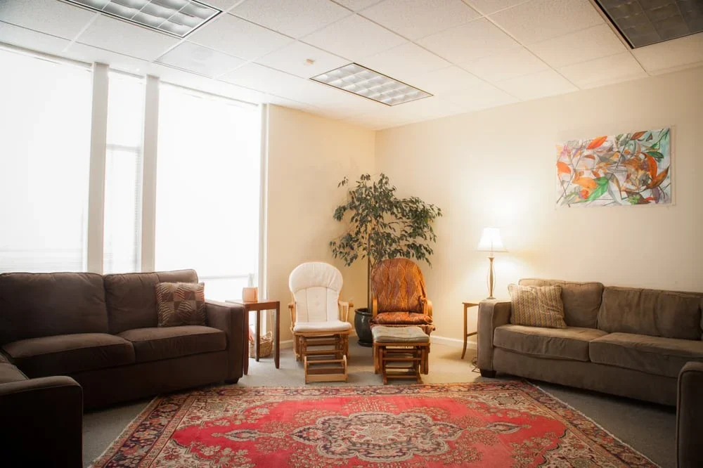Living room with beige walls, a red patterned rug, two beige sofas, two wooden chairs, a white armchair, a potted plant, a table lamp, and artwork on the wall.