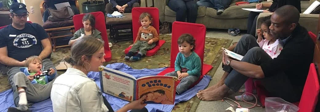 A woman reading a children's book to a group of young children and adults in a cozy indoor setting.