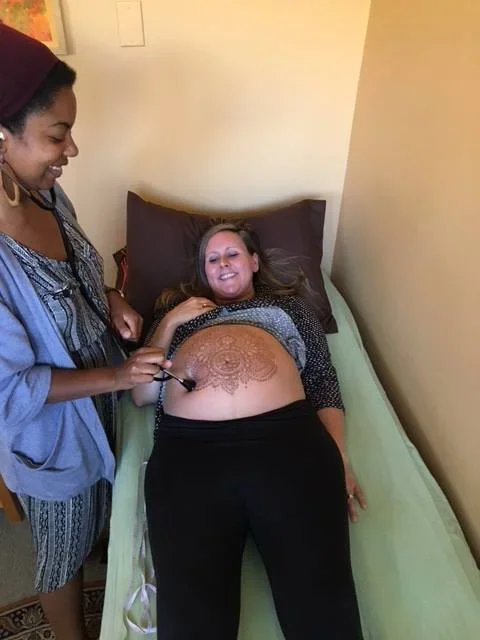 A woman lying in bed while another woman paints a henna design on her belly.