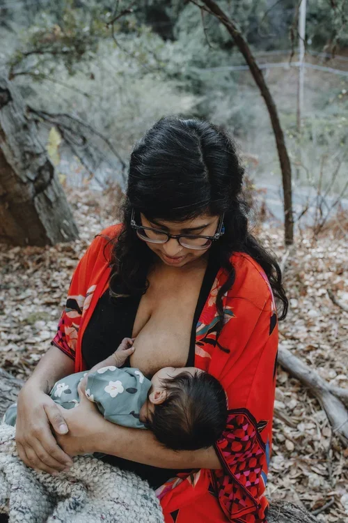 A woman breastfeeding a baby outdoors in a wooded area, wearing glasses and a colorful red and black kimono-style garment.