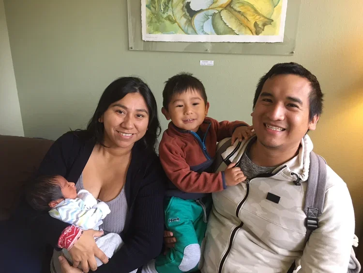 A family of four sitting together on a couch, with a woman holding a newborn baby, a young boy sitting on a man's lap, and the man smiling at the camera. They are in a cozy room with green walls and artwork hanging on the wall behind them.