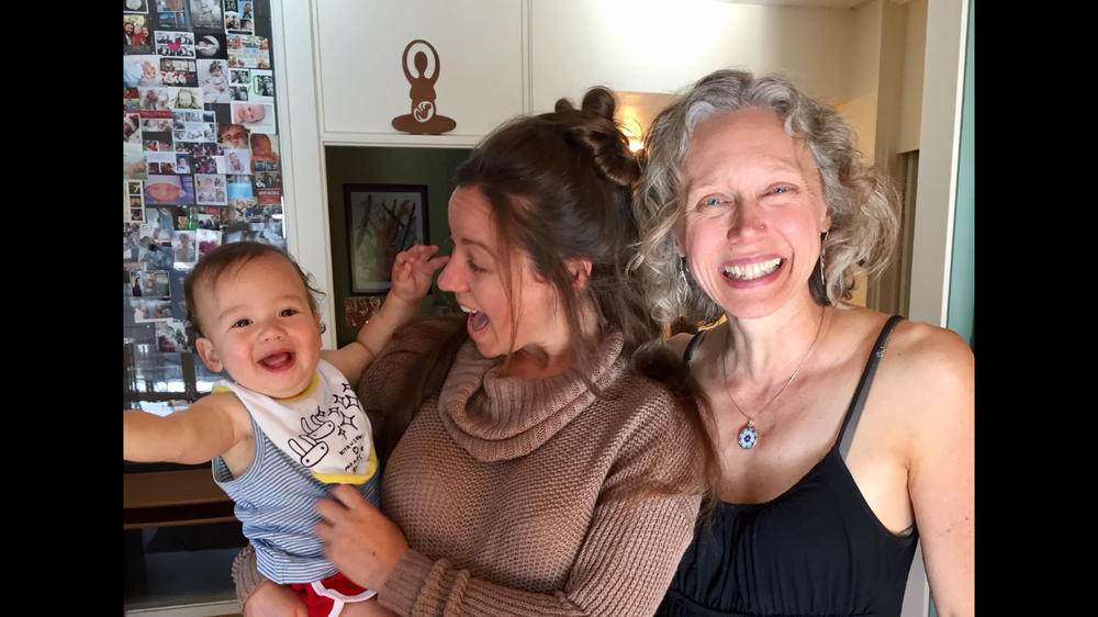 Three people, a baby with a big smile, a woman with brown hair in a bun also smiling, and a woman with curly gray hair smiling, standing indoors in front of a collage of photos and a small wall decoration.