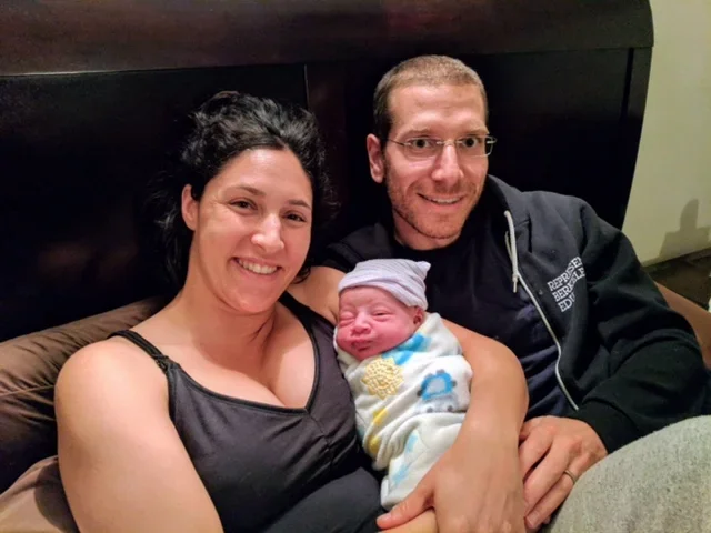 A family of three, including a woman, a man, and a newborn baby, sitting together on a bed. The woman is smiling and wearing a black top, the man is smiling and wearing glasses and a dark hoodie, and the baby is wrapped in a blanket with a hat, showi