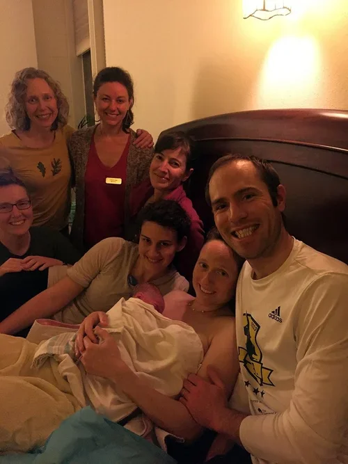 A group of seven people gathered in a room, celebrating childbirth. The woman in the center has just delivered a baby and is holding the newborn, surrounded by family members. Everyone appears happy and excited.