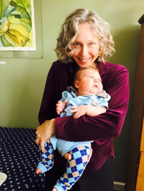 A woman with curly gray hair is smiling while holding a baby dressed in blue and patterned leggings. They are indoors, standing next to a bed with a black and white blanket, with a painting on the wall behind them.