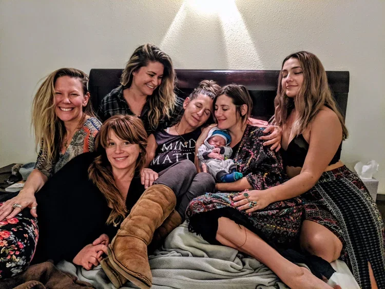 Six women and a newborn baby sitting on a bed, smiling and cuddling together in a cozy room with a soft light above.