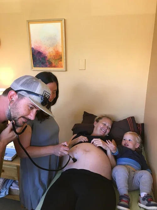 A woman lying on a bed with a happy expression, a young child sitting next to her, and two adults examining her pregnant belly with a stethoscope.