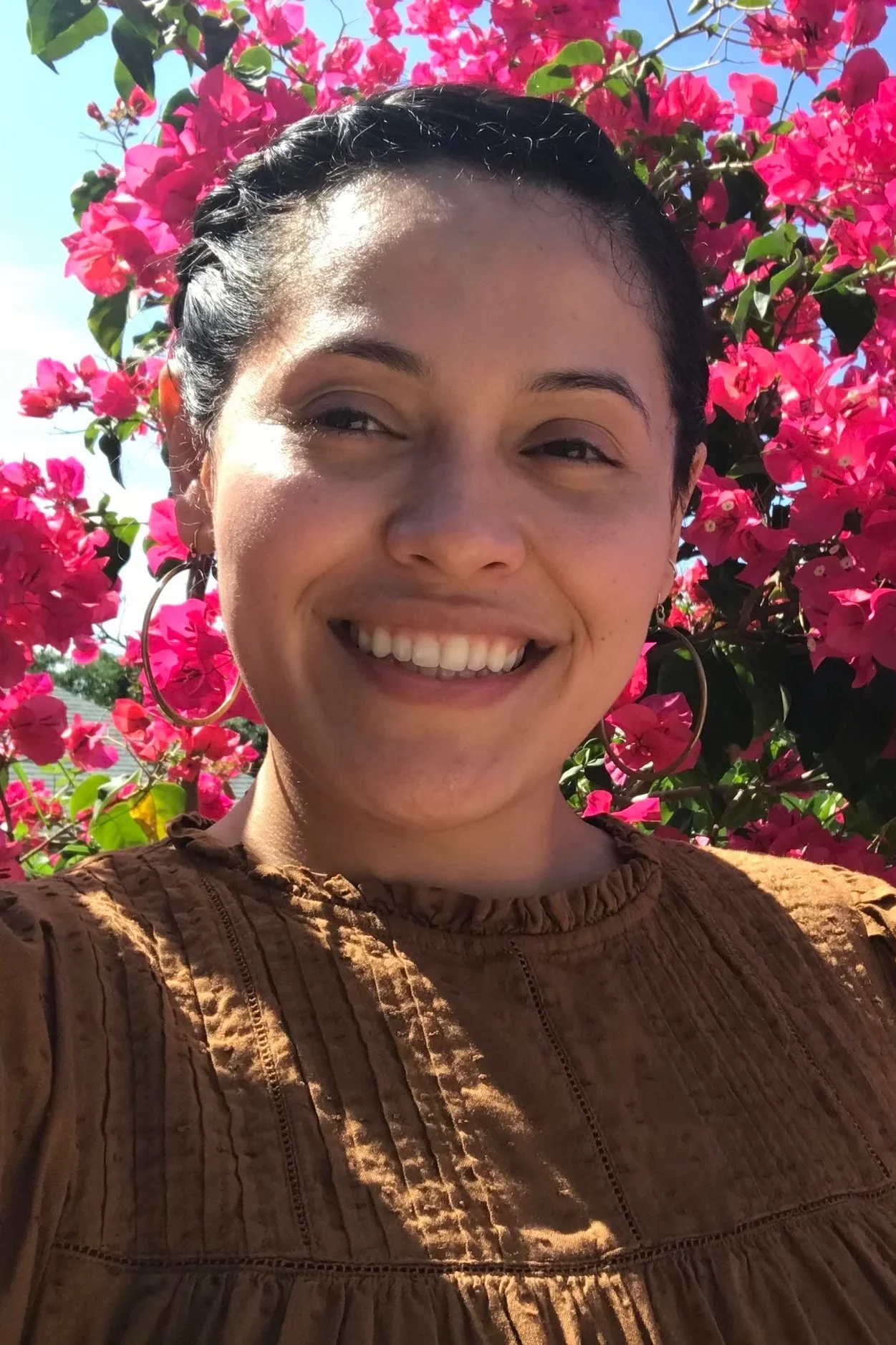 A young woman smiling outdoors in front of a flowering pink bush, wearing hoop earrings and a brown top.