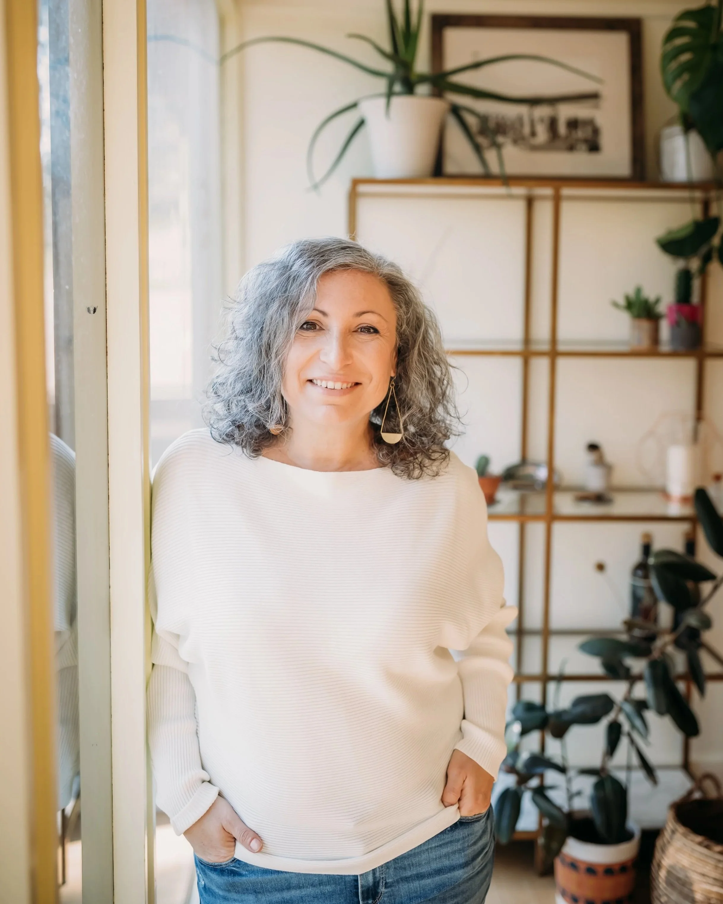 A woman with shoulder-length curly gray hair smiling indoors, wearing a white sweater and blue jeans, standing near a window with a Plant on a shelf behind her.
