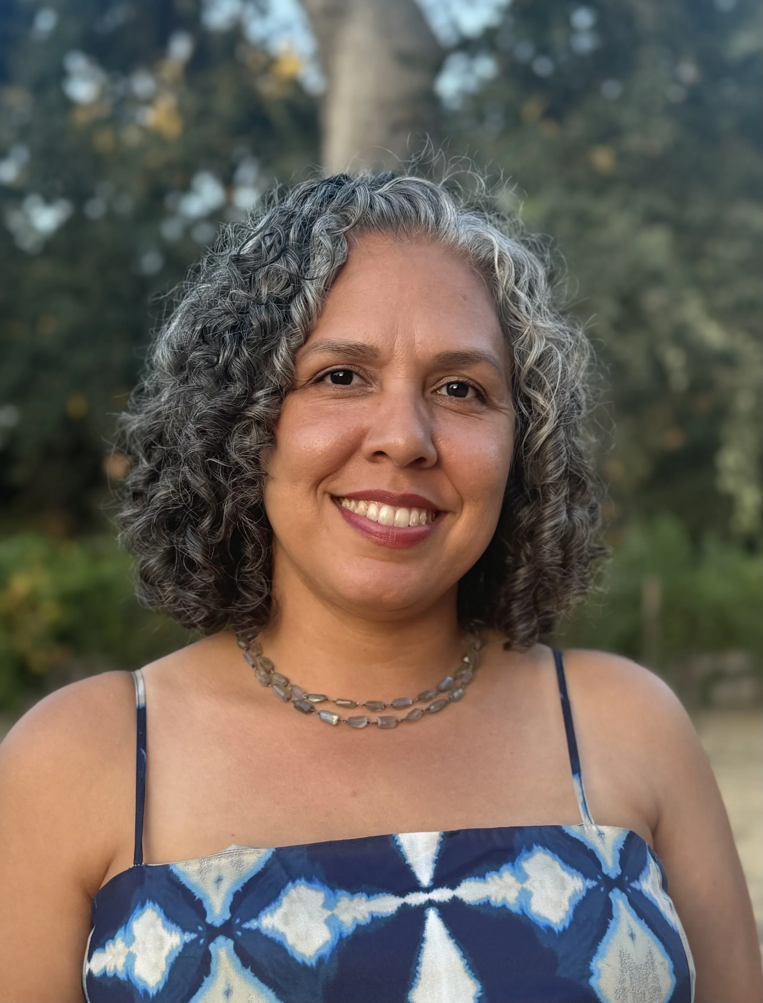 A woman with curly gray hair smiling outdoors, wearing a blue patterned dress and a necklace.
