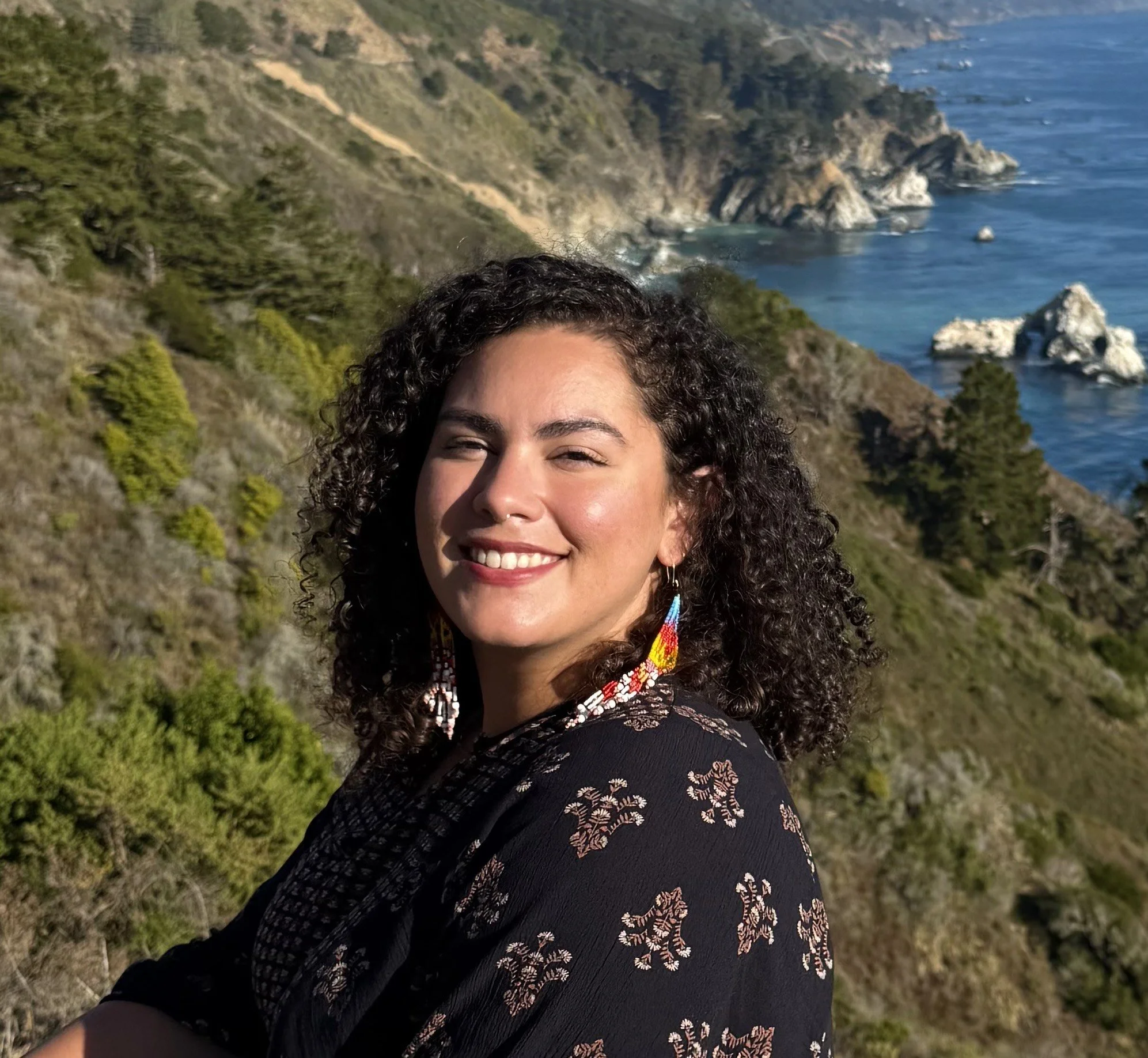 A woman with curly dark hair and colorful earrings smiling outdoors with a coastal hillside and ocean in the background.