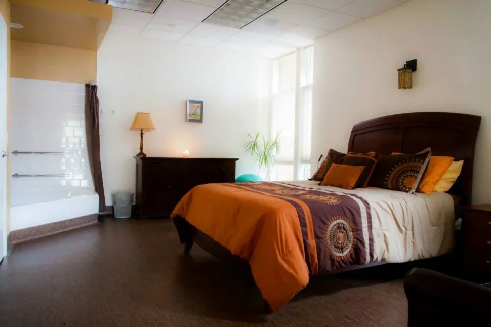 A bedroom with a wooden bed frame, colorful bedding, and multiple pillows, a dresser with a lamp and small decoration, a potted plant, and a window with natural light.