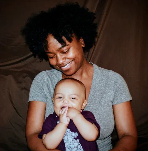 A woman with natural curly hair smiling and holding a young child who is covering their mouth with their hands.